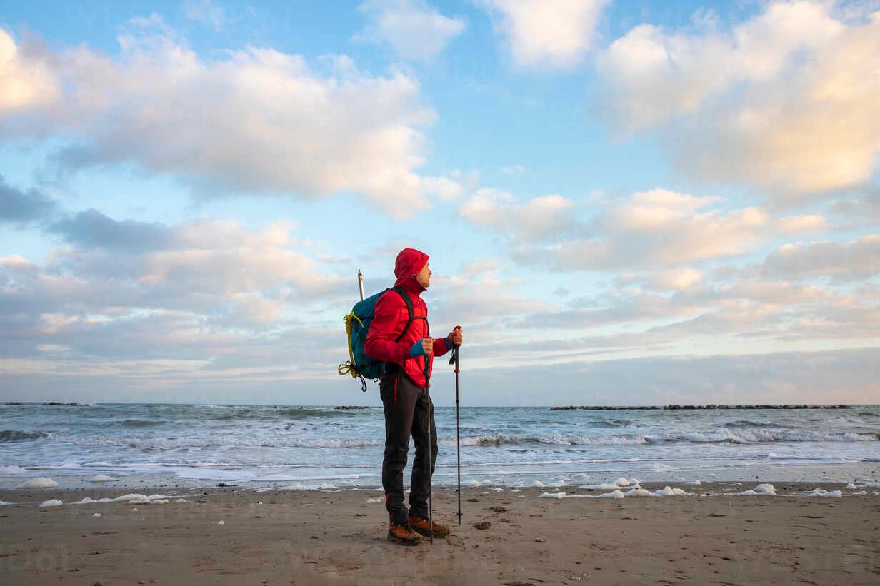 man-with-backpack-and-hiking-poles-standing-at-beach-mediterranean-sea-italy-MCVF00707